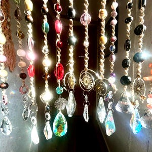 May include: A collection of hanging suncatchers featuring strands of natural jute twine adorned with various colorful glass beads, polished stones, and metallic sun-shaped charms, each ending in a faceted clear crystal prism that reflects light.