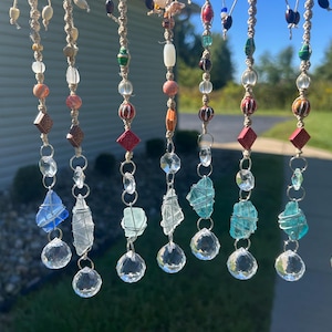 May include: A collection of eight hanging ornaments featuring macrame cords adorned with various colorful beads, including red, white, blue, and green tones. Each strand terminates in a wire-wrapped raw stone and a clear faceted crystal prism.
