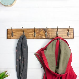 Puede incluir: Un perchero de madera con seis ganchos, teñido de un color marrón oscuro. Una chaqueta roja y una bufanda gris están colgadas de los ganchos.