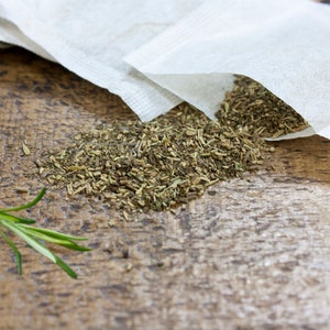May include: A pile of dried herbs, likely a blend, spills out of a white tea bag onto a wooden surface. A sprig of rosemary sits to the left of the herbs.