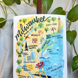 May include: A colorful, hand-painted map of Milwaukee, Wisconsin, featuring landmarks like the MKE Art Museum, Cathedral Square, and Summerfest. The map includes the text "Milwaukee" in a cursive font, with illustrations of a deer, coffee cup, and sailboats.