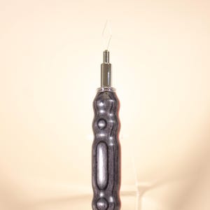 May include: A close-up of a tool with a silver metal tip and a gray and black wooden handle. The handle has a series of rounded, carved sections. The tool is designed for a specific craft or hobby.