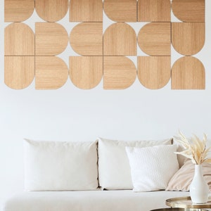 May include: A white sofa with two white cushions and a beige cushion sits in front of a white wall with a geometric wall art piece made of wood. The wall art is made of 16 pieces of wood in different shapes and sizes. The wood is a light brown colour. There are two gold metal side tables with glass tops in front of the sofa. A woven straw footstool sits in front of the sofa.