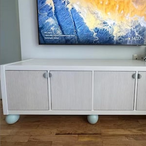 Può includere: Un mobile TV bianco con quattro ante dal design a coste. Il mobile ha piedini sferici azzurri e maniglie argentate. Un grande televisore a schermo piatto è montato sopra, mostrando un'immagine di paesaggio.