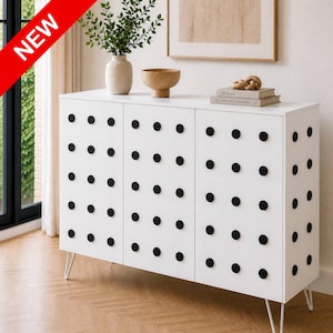 May include: White cabinet with three doors and black circular accents. The cabinet has thin, white metal legs and is topped with decorative items, including a vase with greenery and a wooden bowl. A red banner in the upper left corner reads "NEW".