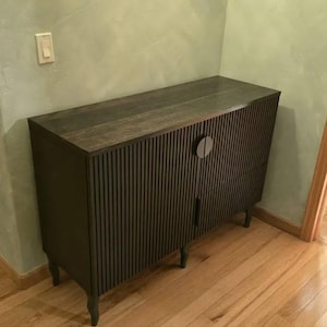 May include: A dark brown wooden cabinet with a ribbed front design and a circular handle. The cabinet has a flat top and sits on four short, curved legs. The cabinet is placed against a light green wall.