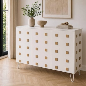 May include: A white cabinet with three doors, featuring square, light brown knobs arranged in a grid pattern. The cabinet has thin, white metal legs. Decorative items, including a vase with greenery and a bowl, sit on top.