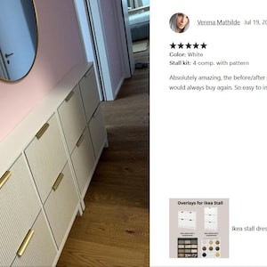 May include: White shoe storage cabinet with gold handles and a textured front panel. The cabinet is mounted on a pink wall, with a gold-framed mirror above. The cabinet has multiple compartments.