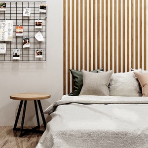 May include: A white bed with a white duvet cover and a brown headboard with vertical wooden slats. There are three pillows on the bed, one green, one beige, and one pink. A small wooden side table with a black metal frame sits next to the bed.