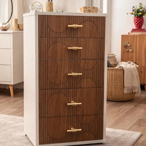 May include: A tall white chest of drawers featuring five drawers with dark wood-toned fronts. Each drawer front displays a geometric pattern of vertical lines and semi-circles, accented by a horizontal gold-toned metal handle in the center.