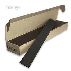 May include: A long, rectangular piece of dark brown Wenge wood with a visible grain pattern, resting against an open, light brown cardboard box. The box features small black shipping icons on the side and the word Wenge printed in white.