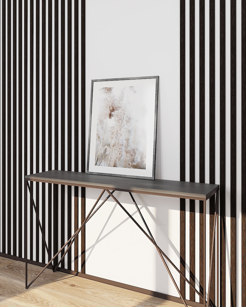 May include: A rectangular picture frame with a thin dark border containing a print of light-colored pampas grass. The frame rests on a slim console table featuring a dark gray top and a geometric metal base with thin, angled support legs.