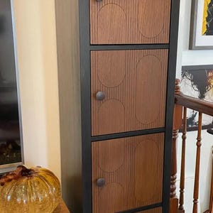 May include: A tall, wooden cabinet with four doors. The doors are a medium brown with vertical lines and oval designs. The cabinet frame is black, and each door has a round, dark knob. A decorative glass pumpkin sits nearby.