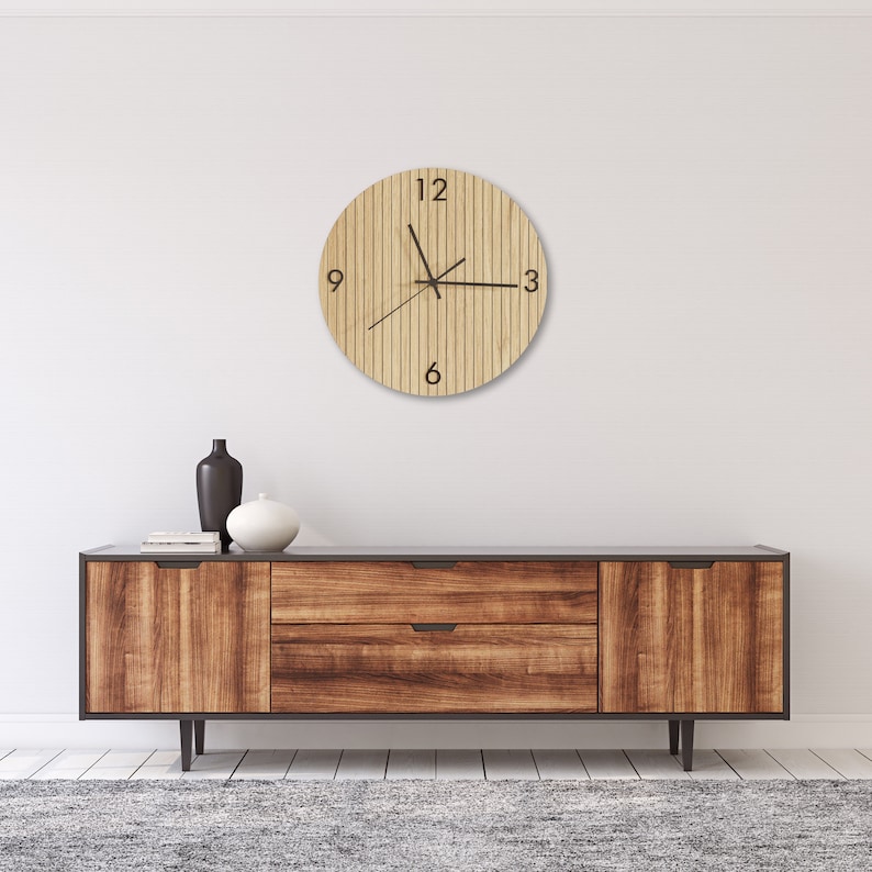 May include: A circular wall clock with a light wood grain finish featuring vertical slats and black numerals at the 3, 6, 9, and 12 o'clock positions. The clock has three thin black hands centered on the face, mounted on a plain white wall.