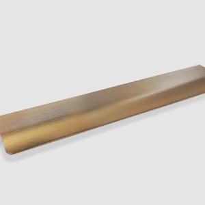 Op de afbeelding: Een lange, rechthoekige meubelgreep van metaal met een geborstelde messing afwerking. De greep heeft een glad, licht gebogen profiel voor randmontage en toont een goudkleurig metalen oppervlak met een subtiele horizontale korrel.