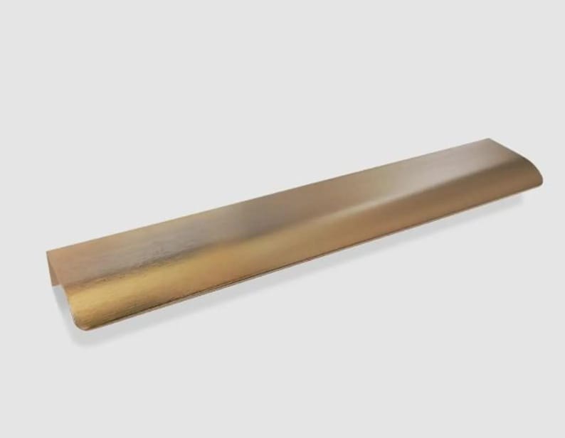 Op de afbeelding: Een lange, rechthoekige meubelgreep van metaal met een geborstelde messing afwerking. De greep heeft een glad, licht gebogen profiel voor randmontage en toont een goudkleurig metalen oppervlak met een subtiele horizontale korrel.