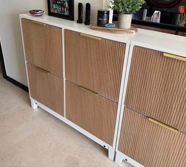 Op de afbeelding: Een witte schoenenkast met drie verticale secties voorzien van lichtbruine, verticaal geribbelde houten panelen. Elke sectie is uitgerust met twee horizontale handgrepen van goudkleurig metaal. De kast staat op korte witte pootjes.