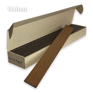 Puede incluir: Una caja de tiras de chapa de madera de nogal. La caja est&aacute; abierta y una sola tira est&aacute; parcialmente sacada. La tira es de color marr&oacute;n claro con un patr&oacute;n de veta de madera.