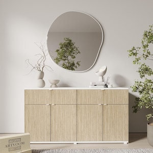 May include: A large, irregular, organic-shaped wall mirror with a thin white frame hangs above a rectangular sideboard featuring four light wood-grain cabinet doors with white trim and small, horizontal, silver-toned metal handles.