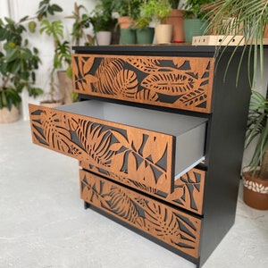 May include: A black dresser with four drawers featuring a tropical leaf design. The drawers are made of wood with a brown stain and have a cutout pattern of leaves. The dresser is standing on a white floor.