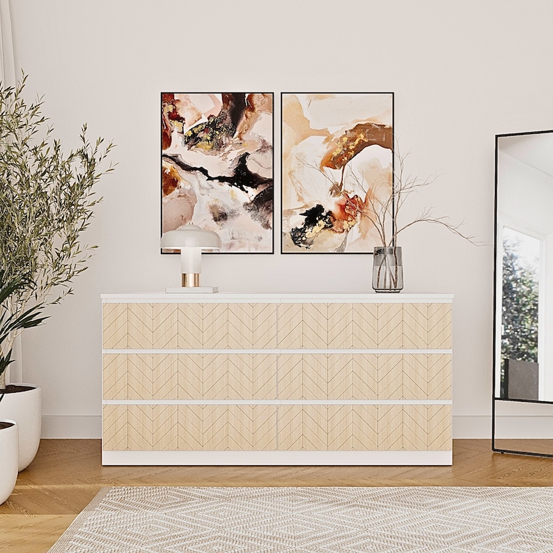 K&ouml;nnte beinhalten: Eine wei&szlig;e Kommode mit drei Schubladen in heller Holzoptik und einem sich wiederholenden Fischgr&auml;tenmuster. Auf der Kommode stehen eine wei&szlig;e Tischlampe und eine Glasvase mit kahlen Zweigen unter zwei gerahmten abstrakten Gem&auml;lden.