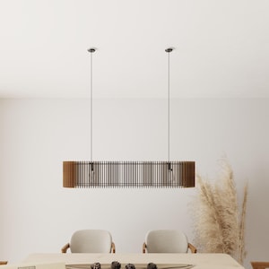 Pode incluir: Uma luminária pendente horizontal com uma estrutura longa de formato oval construída com ripas de madeira verticais na cor marrom escuro. A luz está suspensa no teto por dois fios pretos finos e contém duas lâmpadas visíveis dentro.
