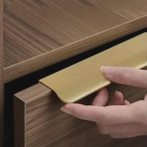 Op de afbeelding: Een goudkleurige metalen randgreep gemonteerd op een ladefront met een houtnerfafwerking. De handgreep heeft een glad, plat oppervlak dat horizontaal over de bovenrand van de lade loopt en een strakke grip biedt voor het openen van de lade.