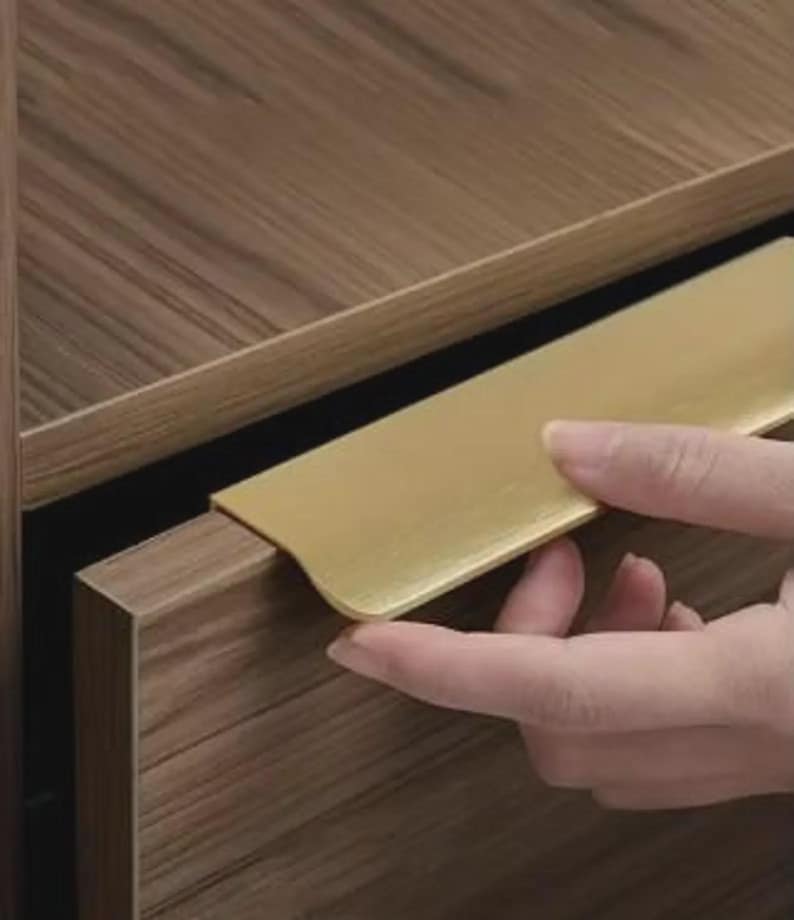 Op de afbeelding: Een goudkleurige metalen randgreep gemonteerd op een ladefront met een houtnerfafwerking. De handgreep heeft een glad, plat oppervlak dat horizontaal over de bovenrand van de lade loopt en een strakke grip biedt voor het openen van de lade.