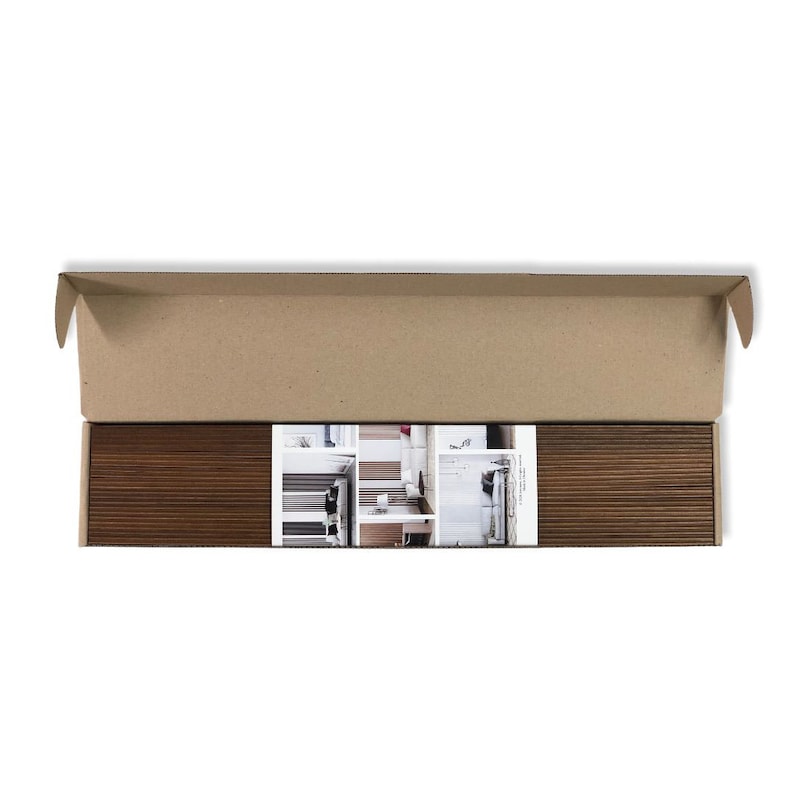 May include: A long, rectangular cardboard shipping box containing two dark brown, horizontally ribbed panels separated by a central white label. The label features a printed photograph of a modern interior space with white walls and furniture.