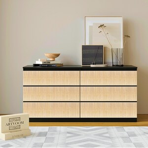 May include: A modern six-drawer dresser with a black top and base, and light wood-toned, vertically-grooved drawer fronts. The dresser is in a room with a neutral color palette, and is decorated with a wooden bowl, books, and a framed print.