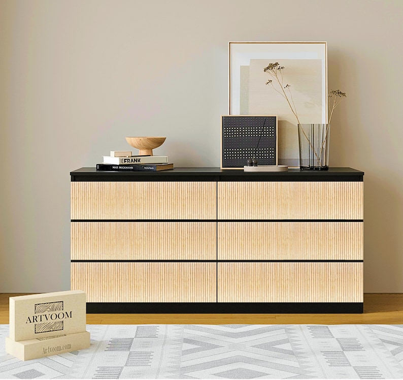 May include: A modern six-drawer dresser with a black top and base, and light wood-toned, vertically-grooved drawer fronts. The dresser is in a room with a neutral color palette, and is decorated with a wooden bowl, books, and a framed print.
