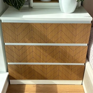 Può includere: Comodino bianco con tre cassetti. Ogni cassetto ha un motivo a chevron in legno di colore marrone e bianco.