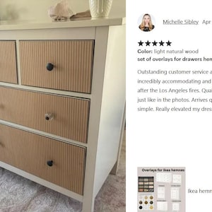Op de afbeelding: Een witte ladekast met acht lades voorzien van licht natuurlijke houtkleurige, verticaal geribbelde overlays. Elke lade is uitgerust met een enkele ronde knop, afwisselend in zwarte en helder glazen afwerkingen op de voorzijde.