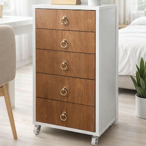 Peut inclure: Un meuble à cinq tiroirs blanc et bois avec des poignées dorées en forme d'anneau. Le meuble repose sur de petites roulettes. Une horloge numérique en bois et une plante en pot sont posées dessus.