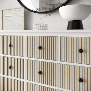Op de afbeelding: Een witte ladekast met zes lades voorzien van verticale lichte houten latten en kleine, ronde, matzwarte knoppen. De ladefronten hebben een gestructureerd, lineair ontwerp en het frame van het meubelstuk is afgewerkt in effen wit.