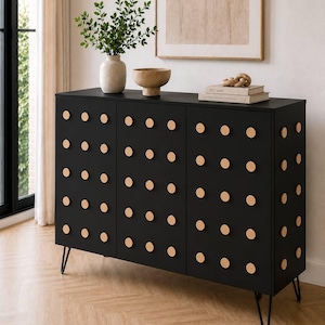 May include: A black cabinet with three doors and numerous round, light-colored wooden knobs. The cabinet sits on thin, black metal legs. A vase with greenery, a bowl, and books are on top. The cabinet is in a room with a window and artwork.