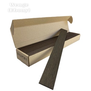 May include: A rectangular cardboard box with an open lid containing several long, thin, dark brown wooden planks with a visible grain pattern. One plank is positioned diagonally in front of the box. The text Wenge (Ebony) is visible above.