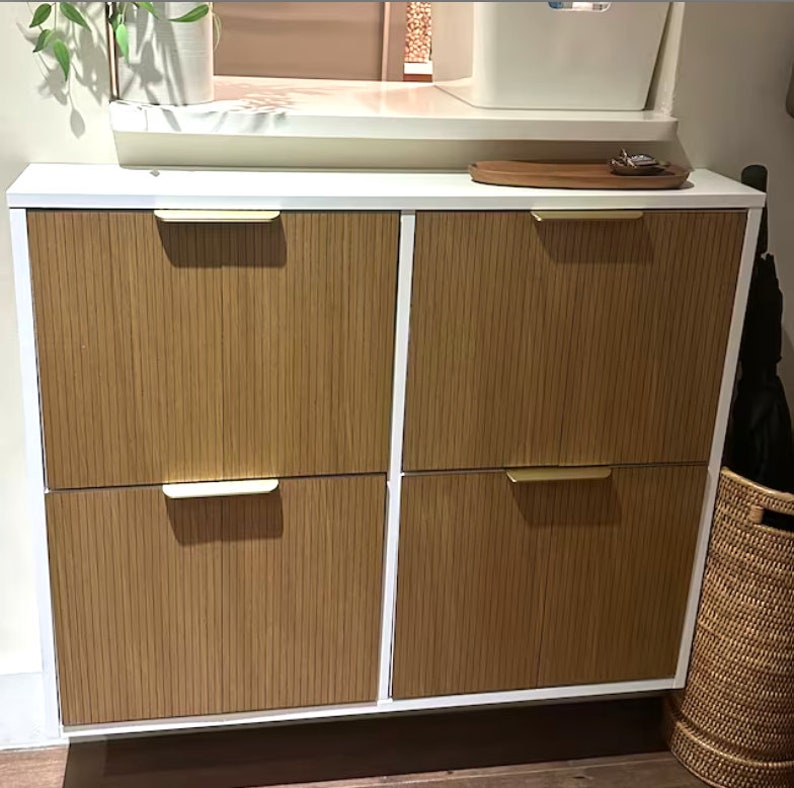 May include: A white cabinet with four doors. The doors are made of vertical wood slats with gold-colored handles. The cabinet is a storage unit, likely for a hallway or entryway. A woven basket is visible on the right side.