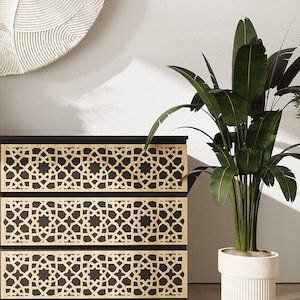 May include: A three-drawer dresser with a black frame and gold geometric pattern on the front. The dresser is next to a potted plant with green leaves in a white planter.