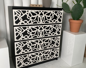 May include: A black dresser with five drawers featuring a white geometric pattern. The dresser is against a white wall with a potted plant and a cactus on a white pedestal to the right.