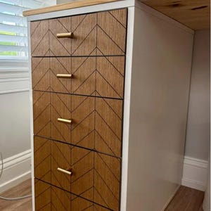 May include: A white desk with five drawers featuring a chevron pattern in shades of brown. Each drawer has a gold-coloured handle. The desk has a light wood top.
