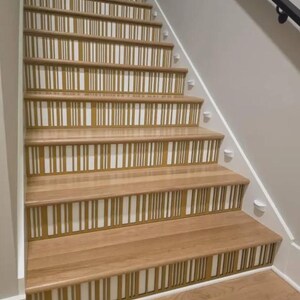 Könnte beinhalten: Holztreppe mit dekorativem Design. Die Stufen bestehen aus hellem Holz mit einem Muster aus vertikalen goldenen und weißen Streifen auf den Setzstufen. Die Wände sind weiß und hellgrau gestrichen.
