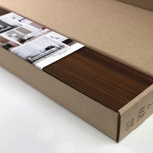 May include: A brown cardboard box with a white label showing images of different rooms with window blinds. The box is open and the inside is filled with a stack of brown wood blinds.