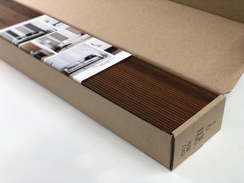 May include: A brown cardboard box with a white label showing images of different rooms with window blinds. The box is open and the inside is filled with a stack of brown wood blinds.