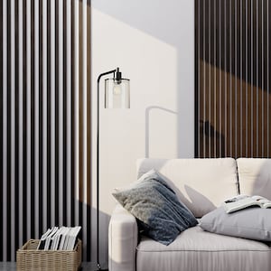 May include: A black metal floor lamp with a clear cylindrical glass shade and an exposed light bulb, positioned next to a light-colored sofa with two throw pillows and an open book, and a woven basket containing several books on the floor.