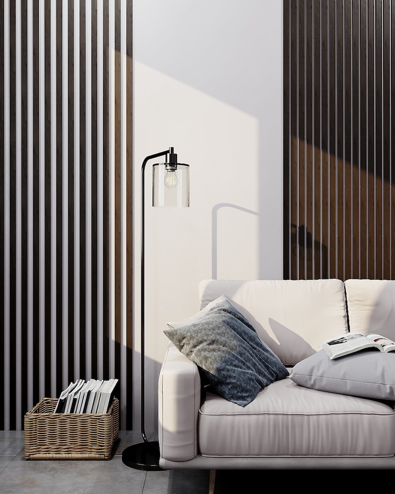 May include: A black metal floor lamp with a clear cylindrical glass shade and an exposed light bulb, positioned next to a light-colored sofa with two throw pillows and an open book, and a woven basket containing several books on the floor.