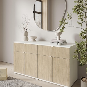 以下が含まれることがあります： 白い天板と明るい木目調のドア、シルバーの取っ手が付いたモダンなサイドボード。有機的な形の鏡が上に設置されています。枝の入った花瓶など、装飾品が白い表面に置かれています。右側には鉢植えの植物があります。