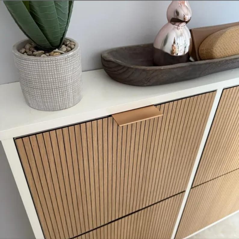 May include: A modern white cabinet with light wood-grain vertical slat doors and a copper-colored handle. A potted succulent and decorative items sit on the white countertop above the cabinet. The cabinet is a stylish storage solution.