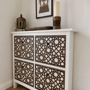 May include: A white cabinet with four drawers, each featuring a dark brown laser-cut geometric lattice overlay. Each drawer has a small black circular knob. The cabinet top holds a metal lantern, a wooden tray, and a tall pillar candle.