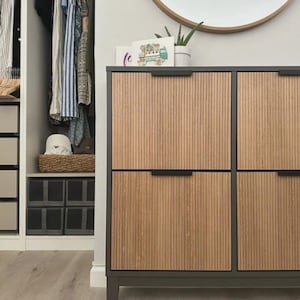 Op de afbeelding: Een rechthoekige opbergkast met een donkergrijs frame en vier lichthouten deuren met verticale latten en dunne zwarte horizontale handgrepen. De kast staat in een kamer met een kledingkast met hangende kleding en opbergbakken.